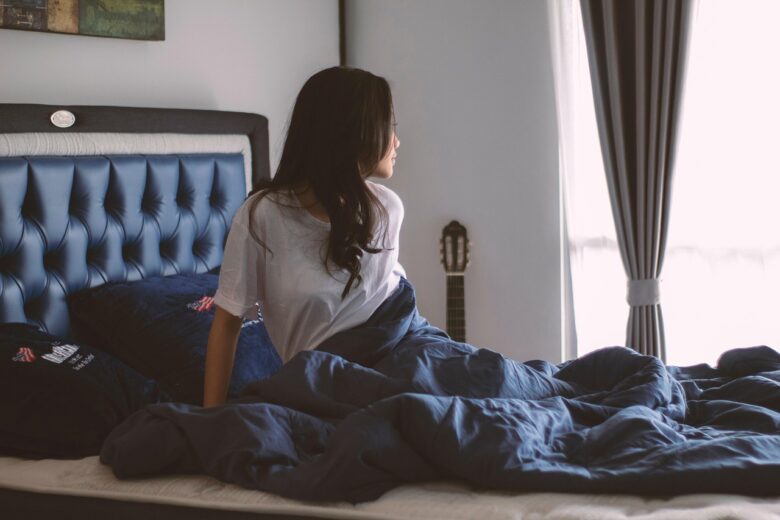 ベッドの上にタオルケットをかけた女性が窓を見ている